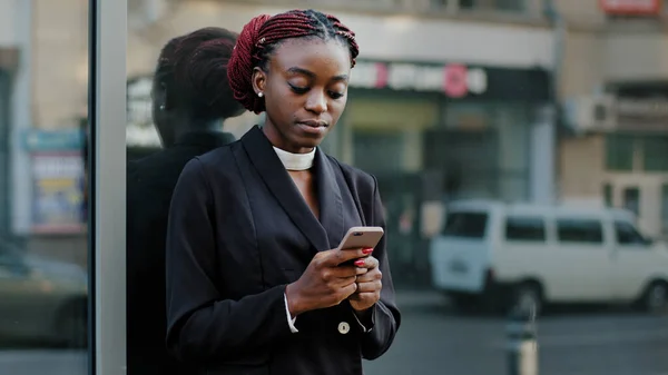 Poważne zaufanie profesjonalny specjalista african amerykański biznes kobieta młoda dziewczyna stoi w mieście w pobliżu budynku biurowego firmy szkła patrzy na ekran telefon komórkowy czat online w smartphone app — Zdjęcie stockowe