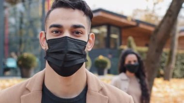 Siyah medikal maskeli çekici esmer adam dikkatle kameraya bakıyor. Şehir parkında ön planda duruyor. Arka planda tanınmayan genç bir kadın.