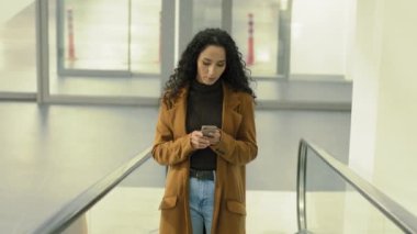 Kendine güvenen alıcı iş kadını cep telefonuna bakıyor akıllı telefondan e-posta okuyor alışveriş merkezinde yürüyen merdivende duruyor. Havaalanındaki merdivenlerde uygulaması olan kadın taksi rezervasyonu.
