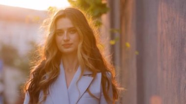 Yakın plan kadın model, uzun kıvırcık sarı saçlı güzel kız güneş ışınları dışında duruyor günbatımının arka planında kameraya bakıyor. Genç hayalperest kadın rüzgarlı havada güneş ışığında poz veriyor.