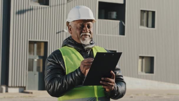 Anciano afroamericano gerente de construcción hombre evalúa edificio viejo capataz de pie en la calle en hardhat protector escribe datos a tableta ingeniero constructor piensa en primer plano plan de trabajo al aire libre — Vídeos de Stock