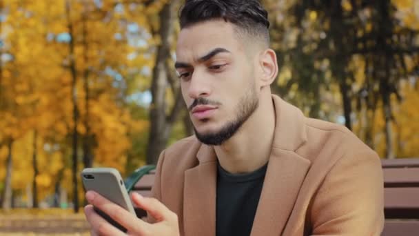 ポートレート若い深刻な男は、携帯電話の画面を見てアプリのパニック男を使用しています電話集中男性のニュースを閲覧秋の公園でベンチに座って読み取りデバイスを使用してビジネスマンが屋外でスマートフォンを保持しています — ストック動画