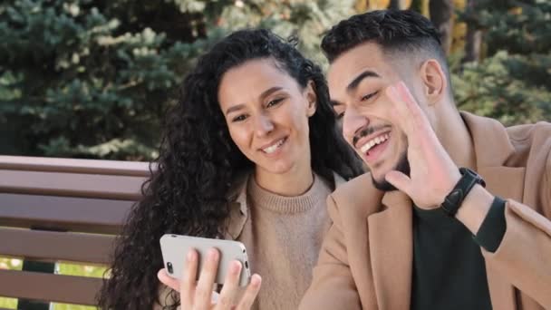 Positive hispanic couple talking online chat use smartphone guy and girl happily smiling waving hello man and woman making video call on device sitting on bench in autumn park joint internet meeting — Stock Video