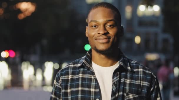 Potret tampan muda african american laki-laki pacar dalam kemeja kotak-kotak berdiri di kota di malam hari melihat kamera tersenyum dengan tulus berpose laki-laki model luar ruangan senyum — Stok Video