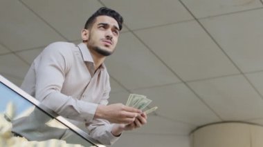 Sonuç olarak, genç, dalgın, sakallı, zengin bir iş adamı elinde bir tomar para tutuyor. Dolar banknotlar, para, finans, kredi, maaş, kredi, kredi yatırımları, balkonda duruyor.