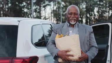 Yakışıklı, yaşlı, Afro-Amerikan bir adam elinde meyve dükkanından alınmış yiyeceklerle gülümsüyor. Kaygısız, emekli, yakın plan erkek. Beyaz arabanın önünde duruyor. Yaşlı bir adam kameraya bakıyor.