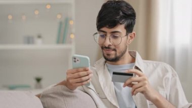İspanyol Arap genç adam, gözlüklü, evde online alışveriş yapıyor internet siparişi veriyor, akıllı telefon ve banka kartı ödemesi yemek dağıtım konseptini kullanarak yemek siparişi veriyor.