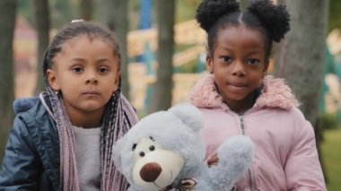 Şehir parkındaki bankta oturan iki arkadaş ellerinde oyuncak ayıyla afro-amerikan kızları gülümseyip okuldan sonra okul çıkışında birlikte oynayan öğrencilere bakıyorlar.