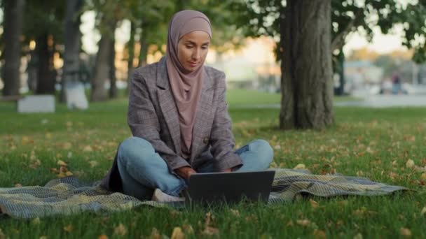 Moslim Arabisch islamitisch student meisje in hijab zakenvrouw freelancer gebruiker maakt gebruik van laptop zittend op groen gras in park werken op afstand studeren in de zomer quarantaine chatten browsen online winkelen — Stockvideo