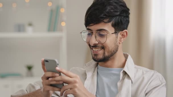 Portrait indoor male arabic guy unshaven man with glasses sitting on couch at home chatting online using mobile phone reads news scrolling browsing net typing in smartphone surprised answers message — Stockvideo