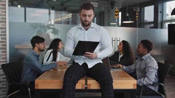 Homem sério sentar na mesa no escritório cheque papelada homem de negócios pensativo com pasta escrever ideias de projeto de negócios diversos colegas multirraciais em segundo plano trabalhando na análise estatísticas financeiras — Vídeo de Stock