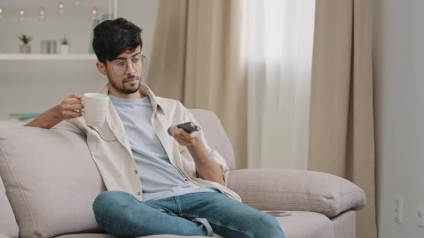 Hombre árabe joven con gafas chico árabe milenario hispano se sienta en el sofá de casa con una taza de café de té cambia canales con control remoto elegir el programa de cine viendo la televisión relajante en el interior fin de semana — Vídeo de stock