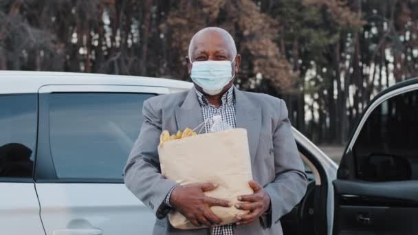 Äldre afrikansk amerikansk man i skyddande medicinsk mask på gatan parkering köpte livsmedel i fruktaffär senior manliga monter nära bil innehar paketprodukter utomhus karantän åtgärder pandemi — Stockvideo