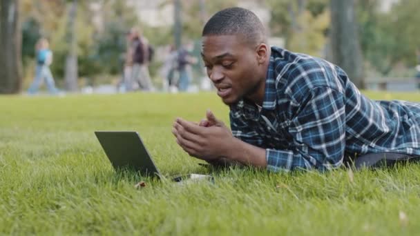 African american guy young business man user lying on green grass lawn in park using laptop online call conference chat remote conversation distant talk talking speaking with friends family outdoors — 图库视频影像