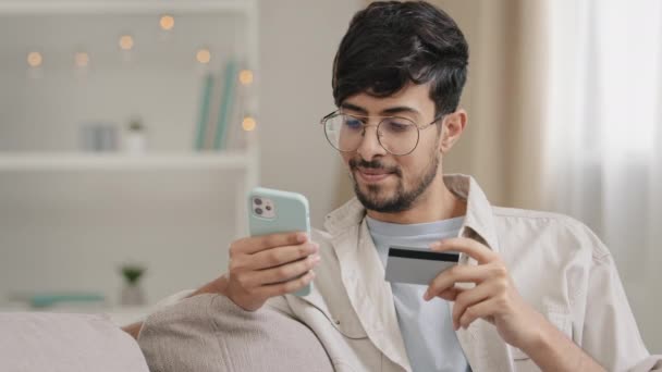 Španěl arabský mladý muž chlap s brýlemi sedí doma nakupování on-line dělá internetovou objednávku lístek objednání potravin pomocí smartphone a bankovní karty čisté platby jídlo dodání finanční koncept — Stock video