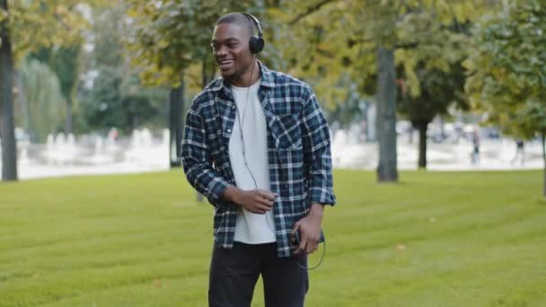 Afroamerykanin stylowy wesoły beztroski człowiek młody szczęśliwy facet w parku nosi słuchawki używa telefonu komórkowego słuchać muzyki korzystając z utworu dźwięk taniec udając gry na perkusji śpiew taniec na zewnątrz — Wideo stockowe