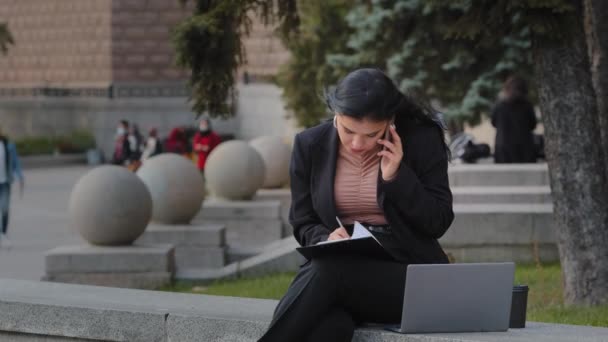 실외에서 일하는 젊은 여성 이 컴퓨터로 온라인 노트북을 가지고 일하는 여성 이 길거리에 앉아 있는 아름다운 히스패닉 여성 이 종이에 자료를 쓰고 있는 것을 발견 했습니다 — 비디오