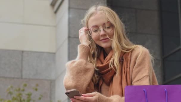 Zakelijke vrouw zit in de buurt van gebouw buiten close-up aantrekkelijk jong meisje communiceert met behulp van telefoon vrouwelijke shopper met pakketten na het winkelen in de stad Kaukasische blonde dame glimlachen typen op gadget — Stockvideo