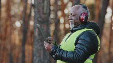 Yaşlı Afro-Amerikan orman mühendisi. Ormanda gürültü yapan kulaklıklar. Yetişkin ustabaşı Foreman, tablet değerlendirmesine veri girerek ağaçları kesme planı üzerinde düşünüyor.