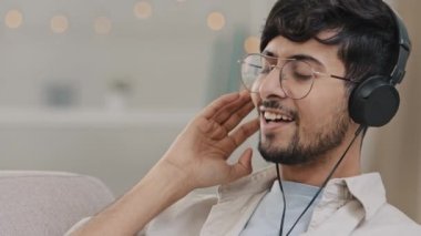 Portre mutlu Arap adam bin yıllık yakın çekim erkek gözlüklü ve kulaklıklı müzik dinliyor evde dinlenerek şarkı söylüyor evde dinleniyor çevrimiçi radyo uygulaması söylüyor