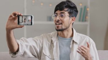 Genç, neşeli, sakallı, gözlüklü adam evde kanepeye oturmuş cep telefonu kamerasıyla video blogu blogcusu Vlogger 'ı kaydediyor ve akıllı telefonuyla internette konuşuyor.