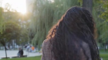 Genç esmer kız, güzel havanın tadını çıkarırken yaz günü parkta gülüyor. Komik portreler çiziyor. Latin, gülümseyen kadın, uzun kıvırcık siyah saçlarıyla doğada duruyor.