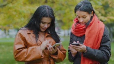 İki genç kadın ellerinin altında telefonla cihazlara bakıyorlar. Biri diğer havalı ücretsiz mobil uygulamaları indirmeye yardım ediyor. İspanyol kızlar sonbahar parkında haftasonu sevgilileri ile çevrimiçi alışverişi tartışıyorlar.