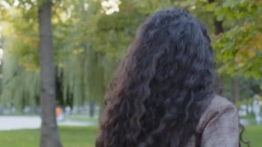 Arka planda hareketli hareketli hareketli esmer, kıvırcık saçlı kız model kız kız öğrenci kız arkadaş zıplıyor kameraya bakıyor, gülüyor, parkta yeşil doğada poz veriyor.