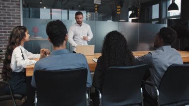 Genç takım lideri, çok ırklı çalışanlara konferans odasında sunum yapıyor. Ofis toplantısında iş adamlarına konuşan kendine güvenen bir erkek, şirket eğitimi projesinde çalışma stratejisini açıklıyor.