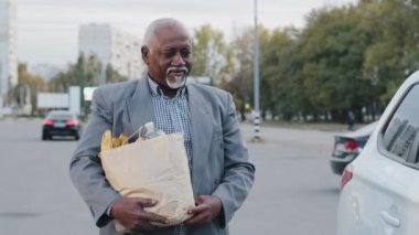 Yaşlı afro-amerikalı beyaz sakallı adam elinde alışveriş çantası taşıyor kaygısız erkek yolcu kapısını açıyor beyaz otomobil işadamı süpermarketten çıkıp açık hava parkına gidiyor arabalara alışveriş yapıyor