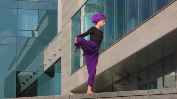 Silny spokojny kobieta muzułmanin islamski dziewczyna w hidżab boso pani w fioletowe spodnie stoi miasto budynku tło zginanie kolano ciągnie biodro do tyłu i z przodu rozciągając jedną nogę równowagi ćwiczyć jogę trening — Wideo stockowe