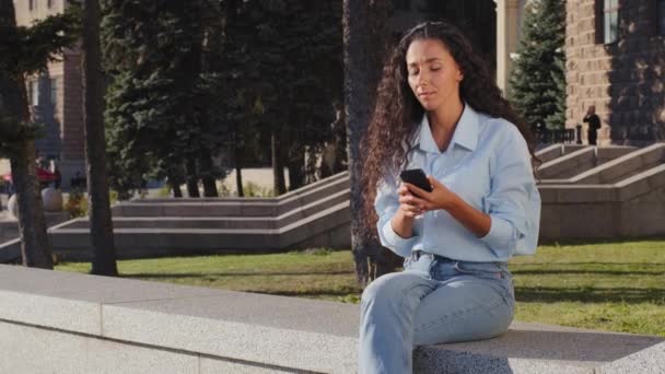Młoda piękna brunetka dziewczyna uśmiecha się czekając na ulicy miasta patrząc na telefon komórkowy czytanie wiadomości machając cześć powitanie gest zaproszenie pochodzą mile widziane symbol przyjazne spotkanie data — Wideo stockowe