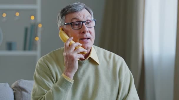 Portrait drôle grand-père à la retraite fait semblant de parler banane comme au téléphone conversation humoristique vieil homme blaguant appelant sur fruit heureux personne âgée souriant joyeusement répondre appel assis à la maison — Video