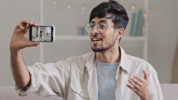 Junge fröhliche arabische Kerl bärtigen Mann mit Brille sitzt zu Hause drinnen Couch mit Handy-Kamera Aufzeichnung Videoblog männlichen Blogger Vlogger winken reden Online-Streaming mit Smartphone — Stockvideo