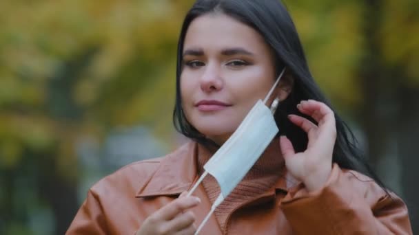 Kapalı alanda kendine güveni olan güzel genç bir kadın tıbbi maskesini takıyor sevimli İspanyol kadın kameraya bakıyor salgın hastalık saçan güzel kız sonbahar parkında duruyor. — Stok video