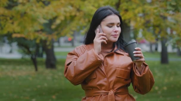 Mladá žena mluví po telefonu se svým přítelem souhlasí setkat veselá krásná dívka odpočívá o víkendu v parku pití lahodné horké kávy s příjemným úsměvem příjemný rozhovor na mobilu — Stock video