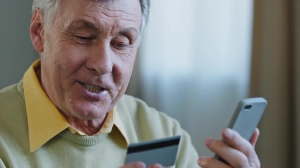 笑顔を閉じる高齢者の男性は、携帯電話の幸せな高齢者を使用してインターネット注文を行いますクレジットカードを保持する高齢者の男性使用オンラインショッピングサービスは、請求書満足クライアントによる電子決済を確保 — ストック動画