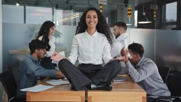 Ung pige meditere i lotus position sidder på bordet i kontor multinationale kolleger skændes i baggrunden smide papirer konflikt på arbejdspladsen attraktiv spansktalende kvinde smilende viser ok – Stock-video