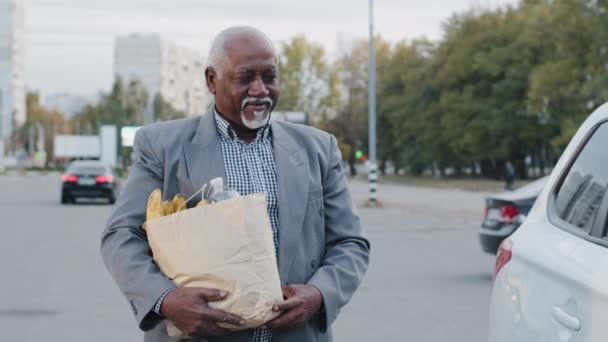 Gammal afrikansk amerikansk man grått hår skägg bär påse med livsmedel i handen bekymmersfri man öppnar passagerardörren vit bil affärsman promenader från snabbköpet utomhus parkering sätter inköp i bil — Stockvideo