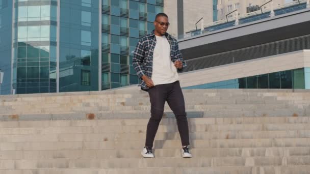 Snygg afrikansk amerikansk man kille modell dansare bär rutig skjorta och solglasögon går uppför trappor i staden utomhus på byggnad bakgrund dans flytta till musik sång dans rörelse promenader gata — Stockvideo