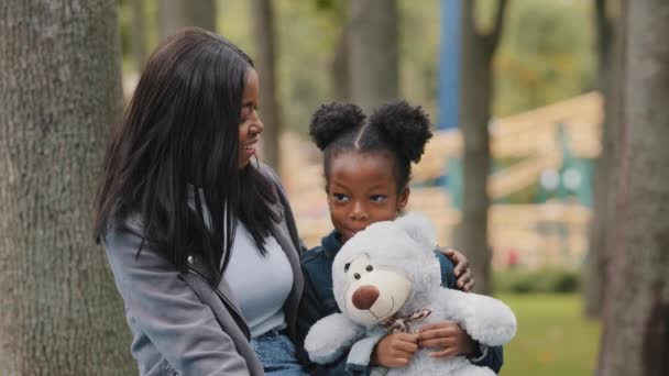 Joyeux maman et fille assis sur le banc dans le parc jeune mère câlins enfant timide petite fille tenant ours en peluche afro-américain famille plaisir à discuter en plein air maman heureux enfant souriant passer du temps avec maman — Video