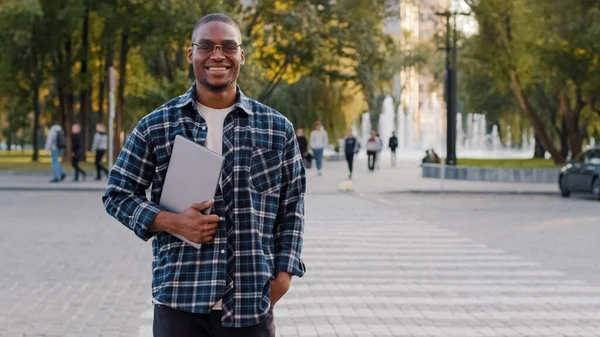 Молодой успешный африканский американец в солнцезащитных очках мужчина в очках бизнесмен с ноутбуком улыбается стоит на перекрестке дорожного движения фоне глядя на камеру позирует на улице улыбается дружелюбно — стоковое фото