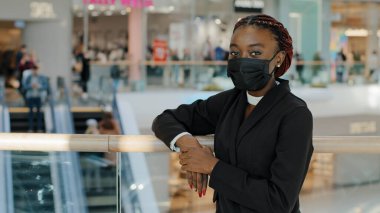 Afro-Amerikalı genç kadın tüketici portresi alışveriş merkezinde dikilip salgın zamanında dükkan yürüyen merdivenin yanında poz veren kameralara bakarken tıbbi maske takıyor.