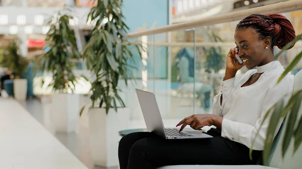 Młoda Afroamerykanka dziewczyna freelance studentka siedzi na kanapie w centrum handlowym biuro firmy rozmawia przez telefon komórkowy odpowiadając na rozmowy wpisując pracując z laptopem pani wielozadaniowość — Zdjęcie stockowe