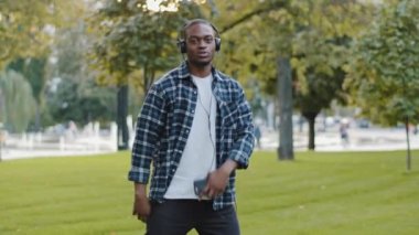 Genç Afro-Amerikalı, kulaklıklı yakışıklı, cep telefonlu, müzik dinleyen, yeşil çimlerin üzerinde parkta dans eden, müzik dinleten yakışıklı bir adam.