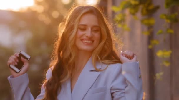 Outdoors female portrait beautiful stylish girl woman lady with long curly hair listens to music with mobile phone online sound app dancing moves in sunlight at sunset in city on street enjoying dance — Stock Video