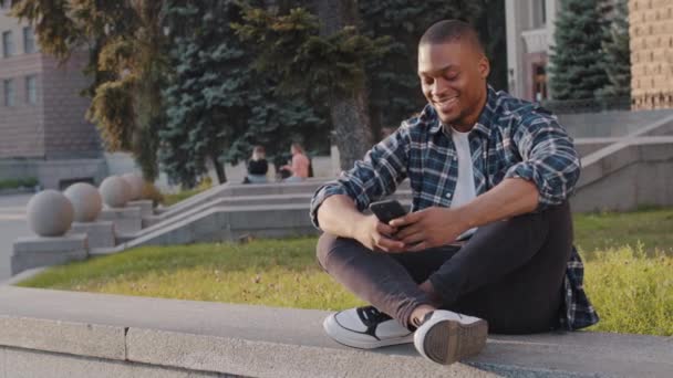 거리에 앉아 야외에 앉아 핸드폰을 보고 있는 행복 한 아프리카 계 미국인 청년은 멀리서 손을 흔들며 인사를 나누고 있습니다. — 비디오