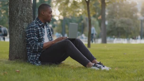 Cansado estudiante chico aburrido hombre de negocios afroamericano hombre freelancer sentado en césped de hierba verde en el parque sensación de dolor de cuello incomodidad cierra portátil dejar de trabajar aprendizaje descansando relajante tomar descanso — Vídeo de stock