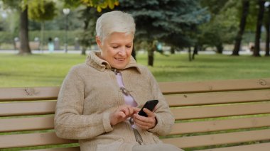 Aşırı neşeli, kır saçlı, dışarıda oturan olgun bir kadın, telefonla akıllı telefon uygulamaları kullanıyor, ekrana bakıyor, son sınıf öğrencileri ile internetten sohbet ediyor, alışveriş yapıyor, mobil cihazlarla eğleniyor.