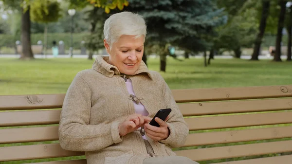 Aşırı neşeli, kır saçlı, dışarıda oturan olgun bir kadın, telefonla akıllı telefon uygulamaları kullanıyor, ekrana bakıyor, son sınıf öğrencileri ile internetten sohbet ediyor, alışveriş yapıyor, mobil cihazlarla eğleniyor.